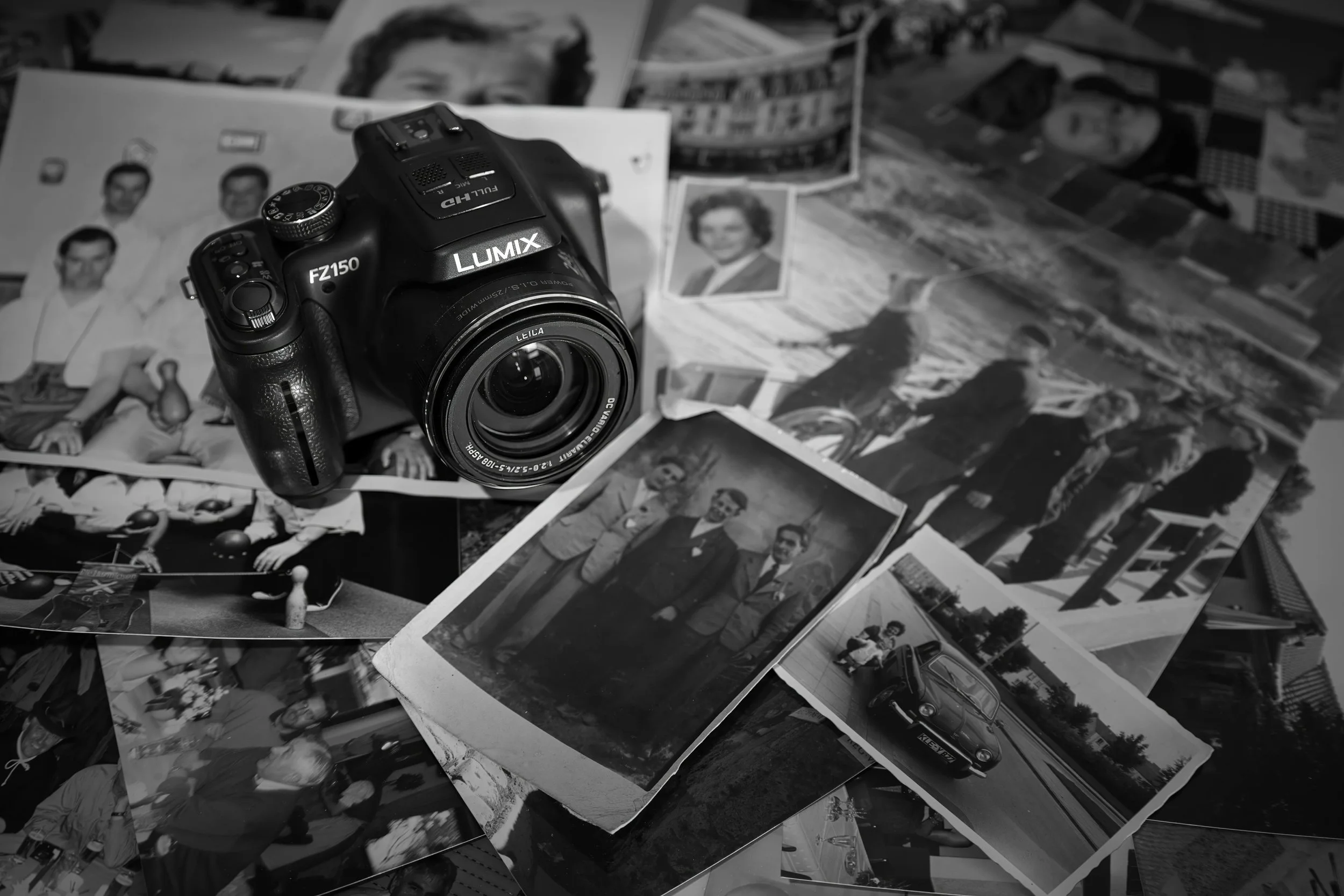 Vintage family photographs scattered on a table with a Lumix camera placed on top, representing reviewing and re-examining old genealogy records.