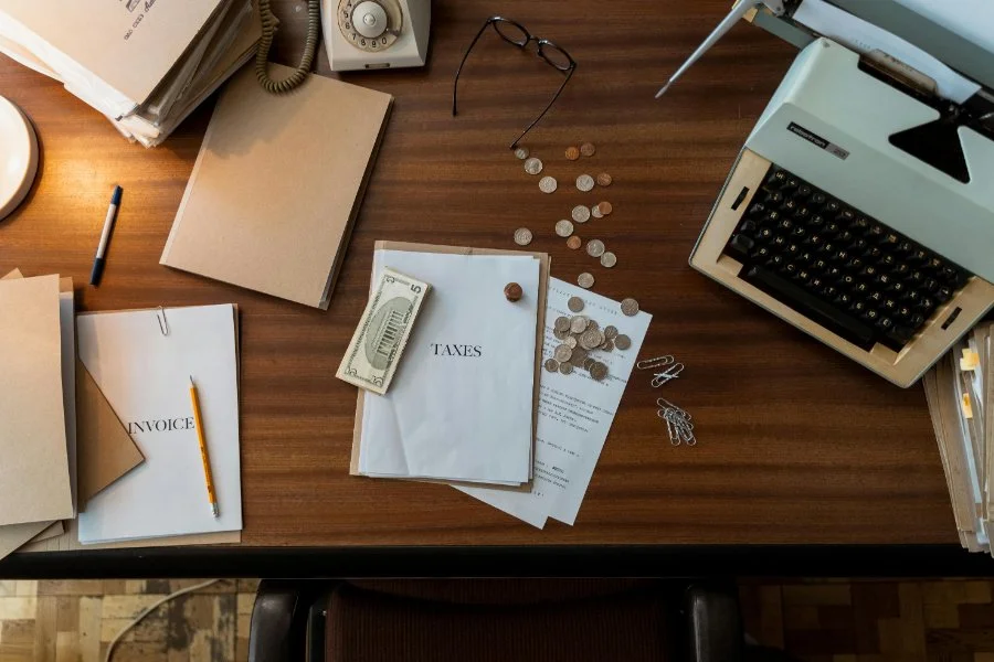 Overhead desk view with papers labeled “TAXES,” coins, and an old typewriter, a workspace for reviewing tax records and family history research.