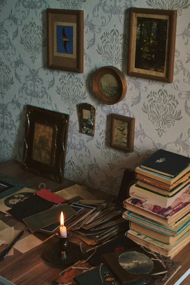 Vintage family photographs, handwritten papers, and stacked journals sit beside a candlelit desk beneath framed artwork and patterned wallpaper. AI and genealogy