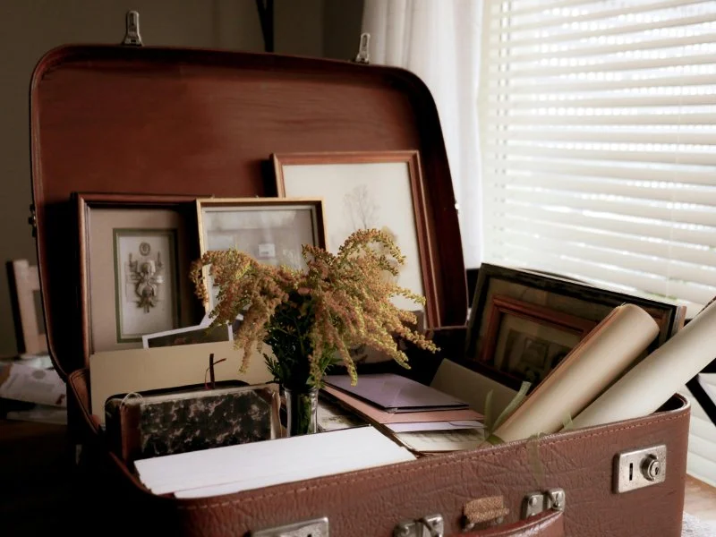 Vintage suitcase filled with framed photos, papers, and keepsakes by a window.