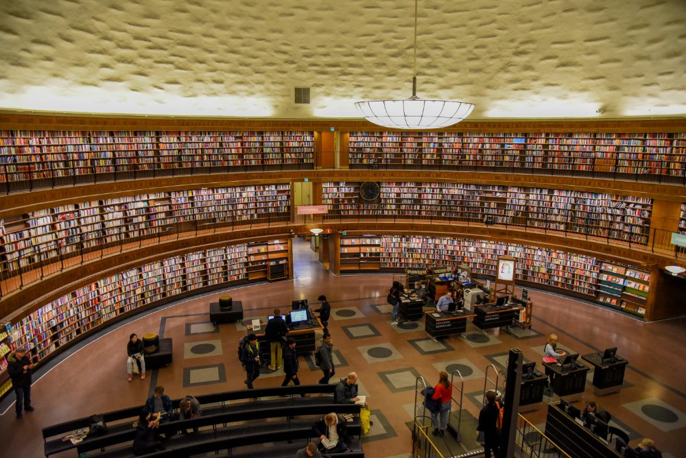 Circular public library reading room with curved walls of bookshelves, people studying at tables, and a large ceiling light, showing types of libraries used for genealogy research.