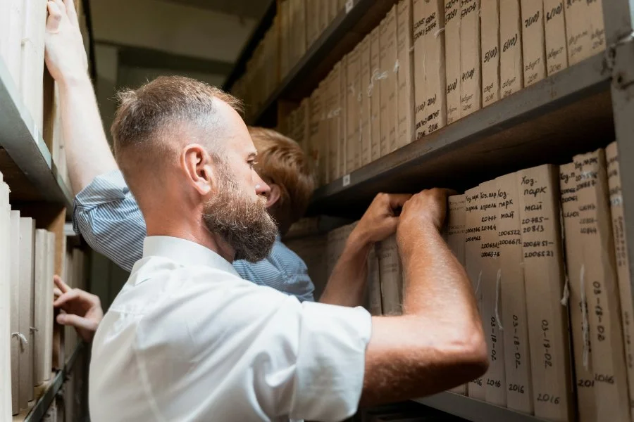 Man and colleague pulling labeled archive boxes from shelves in a records room, showing how traditional research supports the best DNA tests by uncovering family history details found only in physical archives.
