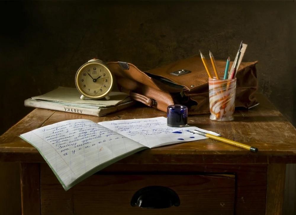 Vintage desk with an open handwritten notebook, pencils, ink, and an alarm clock, like a genealogy research log setup for recording sources.