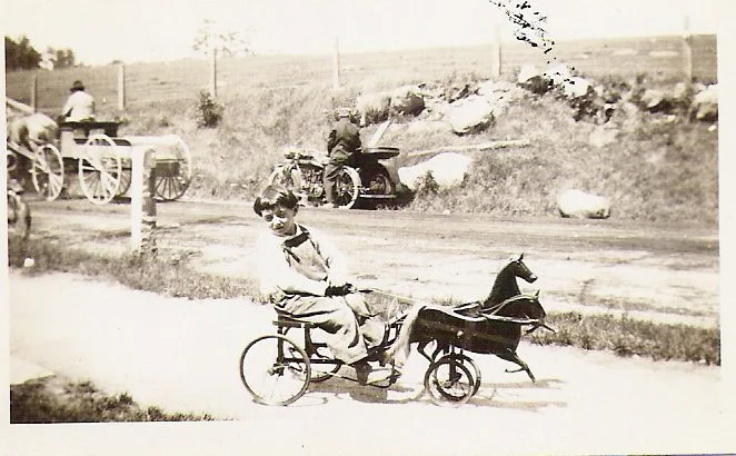 Antique photo of a young boy on toy carriage with men on a horse carriage and motorcycle in the background