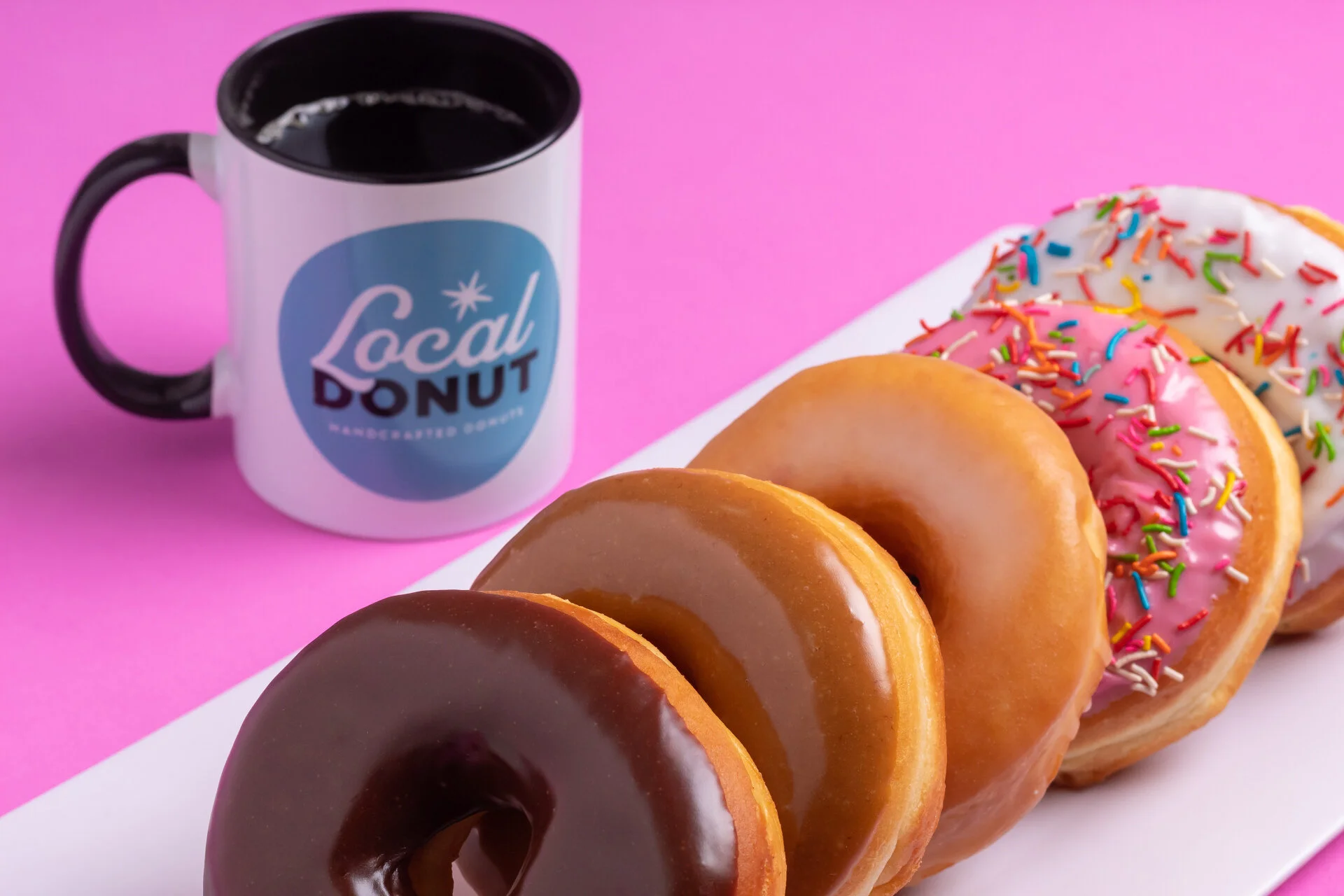 Taza de café con logo de 'Local Donut' junto a cuatro donas de diferentes sabores y glaseados con chispas de colores en una bandeja blanca sobre fondo rosa.