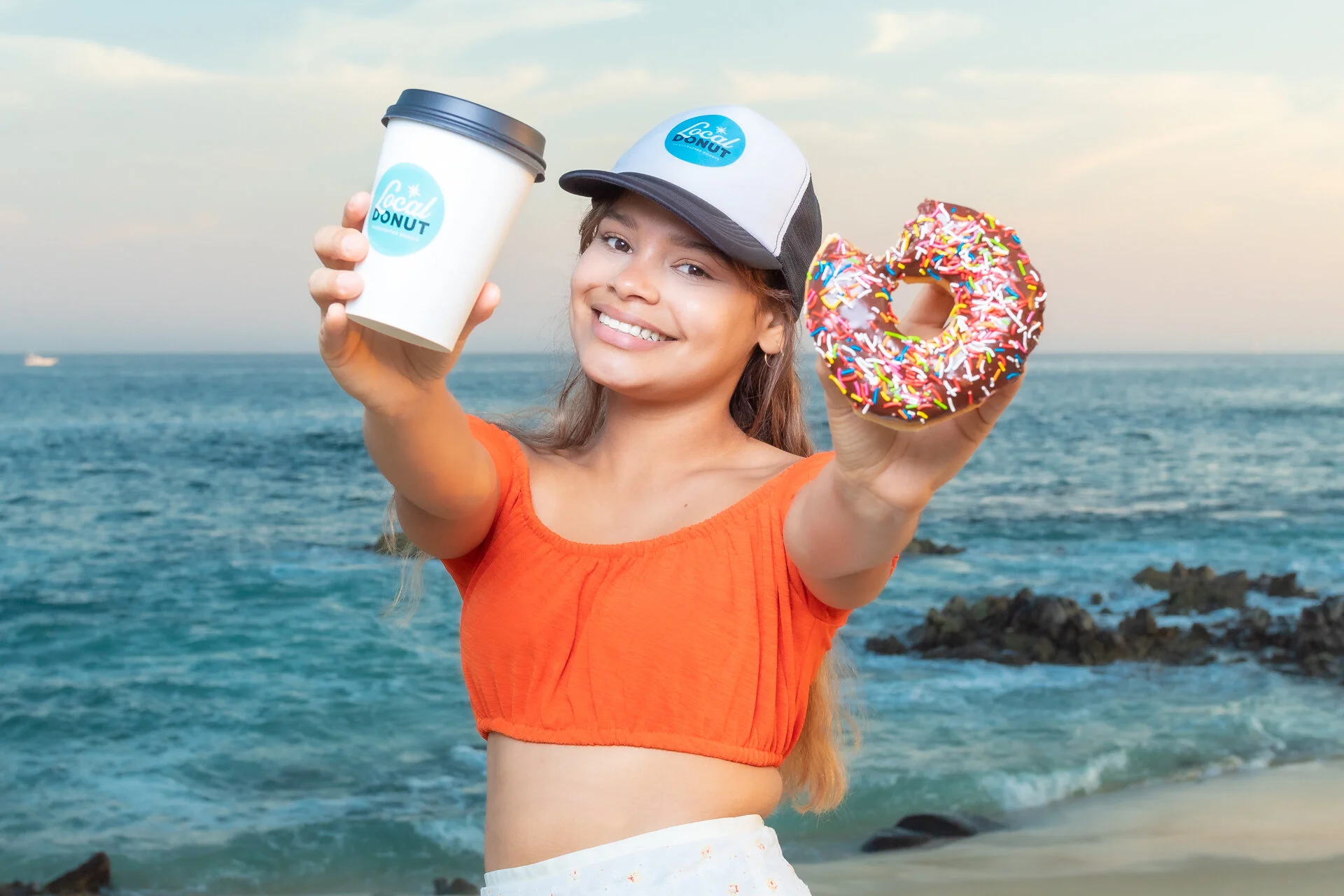 Joven sonriendo en la playa, sosteniendo un café para llevar y un donut con glaseado de chocolate y chispas coloridas.