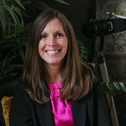 A woman with long brown hair, wearing a black blazer and a pink blouse, smiling in an indoor setting with green plants and a camera tripod in the background.