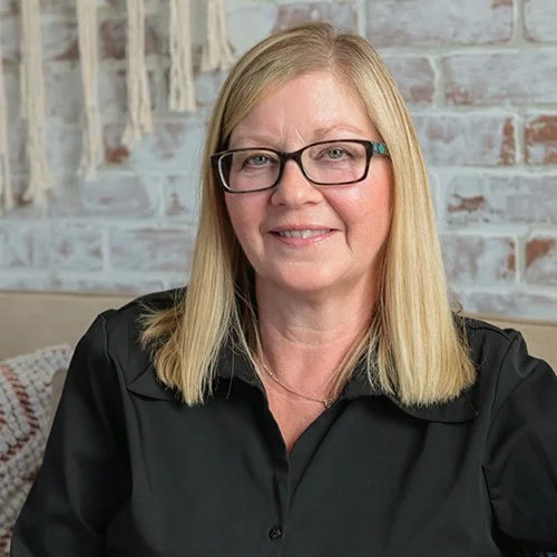A woman with blonde hair, glasses, and a black shirt sitting in front of a brick wall.