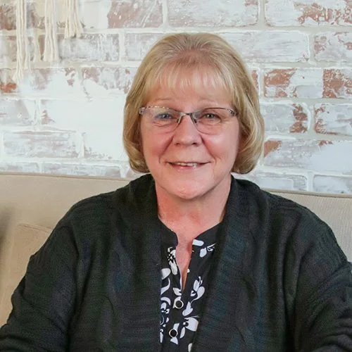 An older woman with blonde hair and glasses, smiling while sitting on a beige couch against a white brick wall.