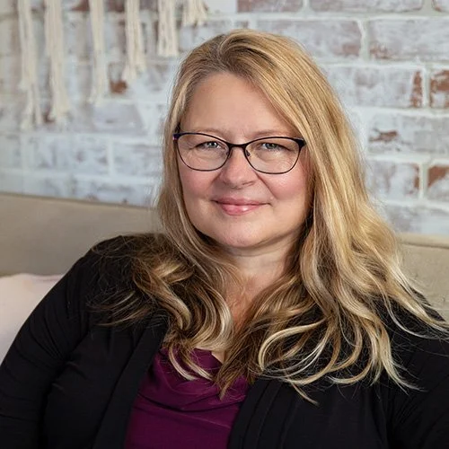 A woman with blonde hair, glasses, and a black cardigan sitting on a beige couch against a brick wall background.