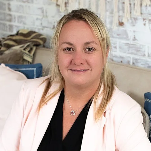 A woman with shoulder-length blonde hair wearing a light pink blazer and a black top, sitting in a cozy room with a brick wall and pillows in the background.