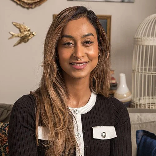 A woman with long, light brown hair smiling at the camera indoors, with a birdcage and decorative items in the background.