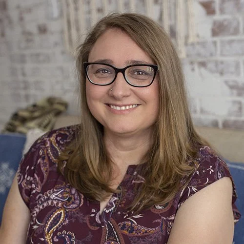 A woman with long brown hair, glasses, and a friendly smile posing indoors against a brick wall.