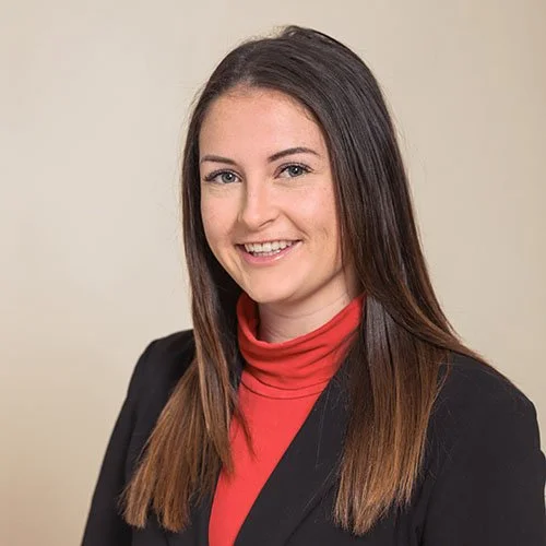 A young woman with long brown hair, wearing a black blazer and red turtleneck, smiling in front of a plain beige background.
