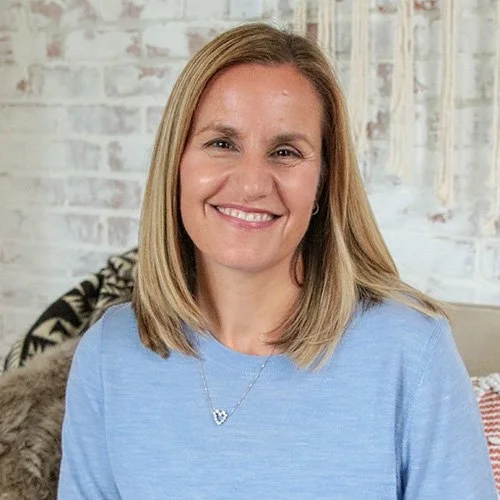 A smiling woman with shoulder-length blonde hair, wearing a light blue top and a heart-shaped necklace, sitting on a beige sofa in a room with a brick wall background.
