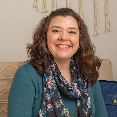Smiling woman with curly hair wearing a teal top and floral scarf, sitting on a couch with wall hanging behind her.