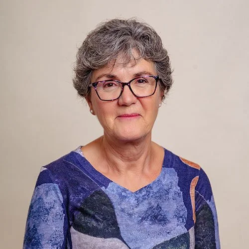 Portrait of an older woman with gray curly hair and glasses, wearing a blue patterned top, standing against a plain light-colored background.