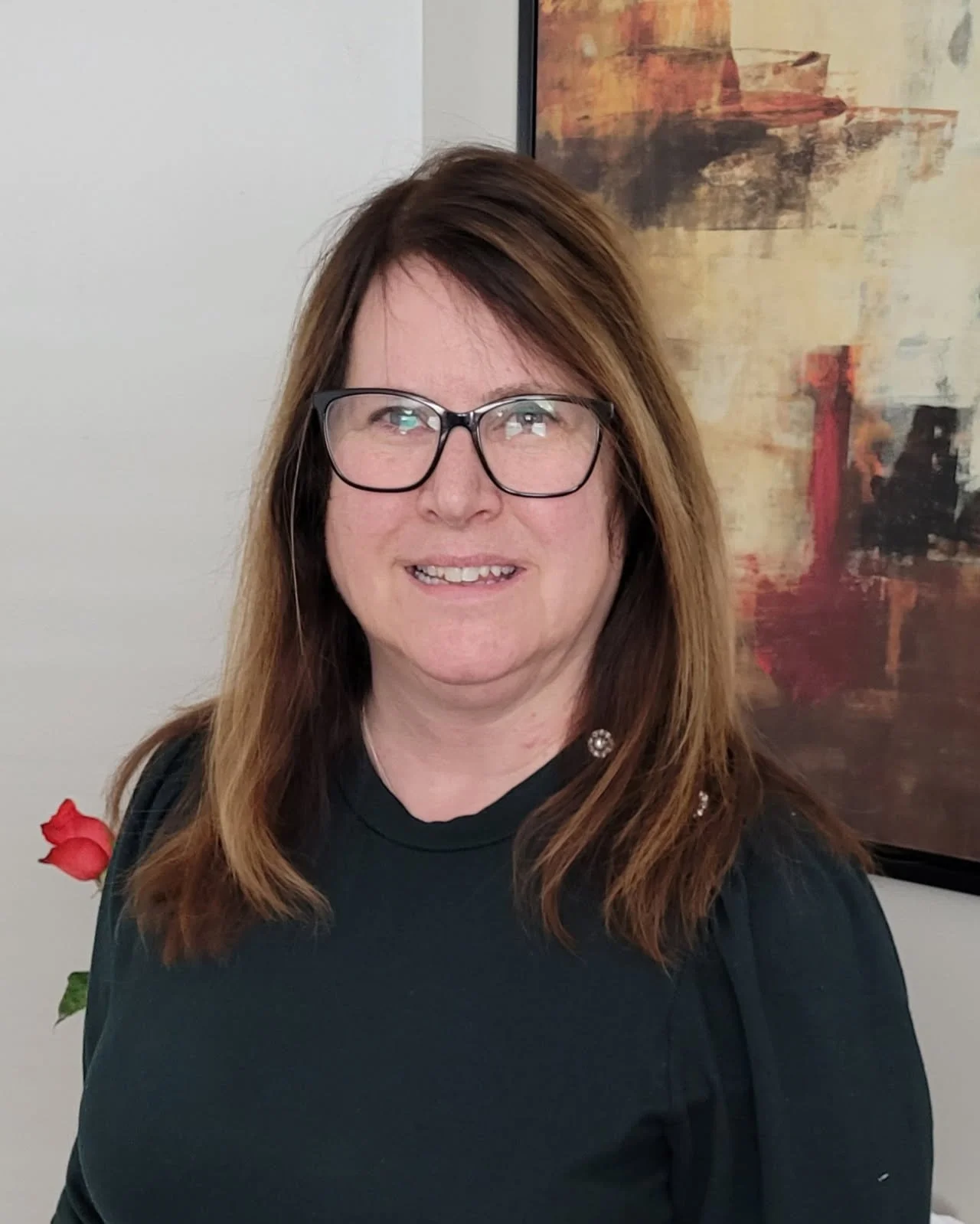 A woman with brown hair and glasses smiling at the camera, standing in front of abstract artwork on a wall with a rose plant to her left.