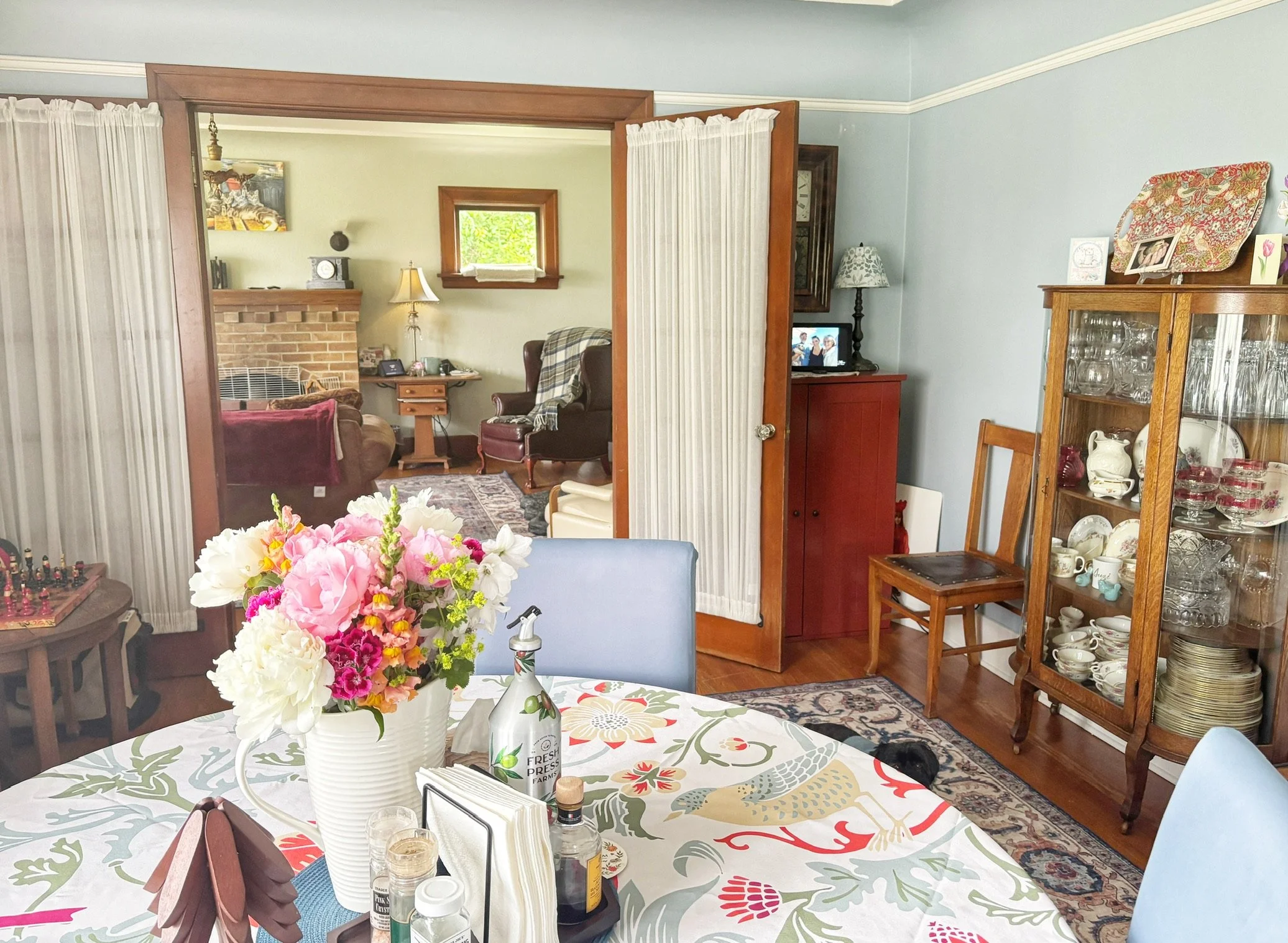 Bright and welcoming dining room with vintage charm, featuring a floral table setting, fresh flowers, hardwood floors, and elegant wood trim opening into a cozy living room with a fireplace.