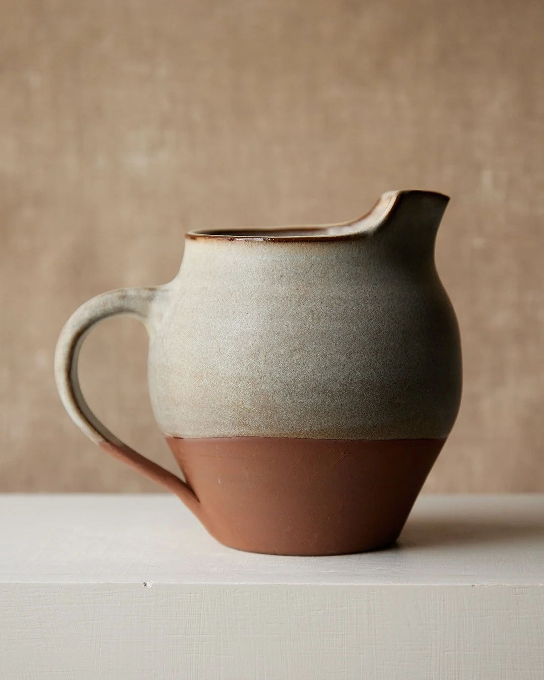 Hand-thrown stoneware pitcher with grey glaze breaking into natural clay in a gentle, organic line.