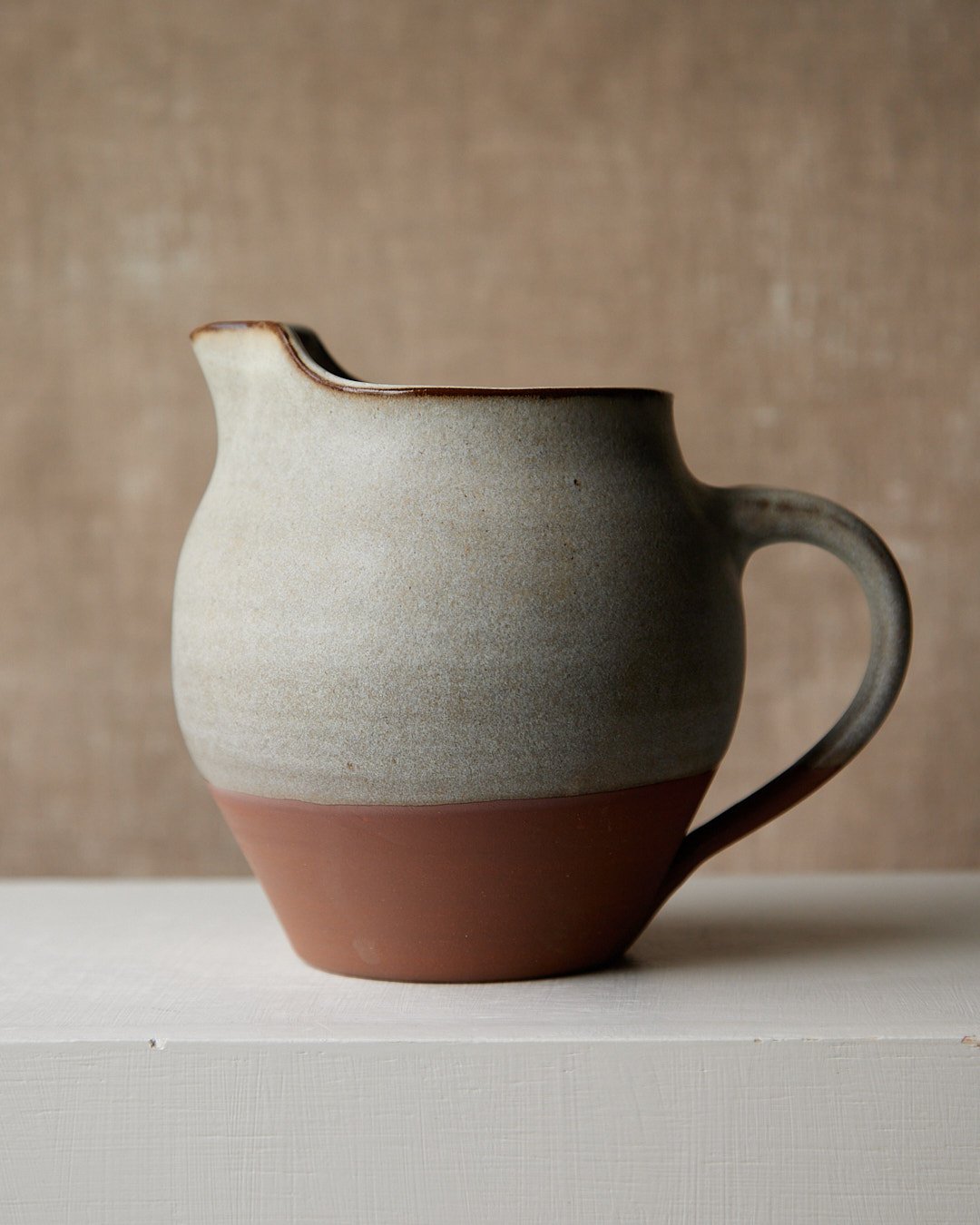Stoneware pitcher with a soft grey glaze flowing into warm clay, rounded body, and raw clay base.