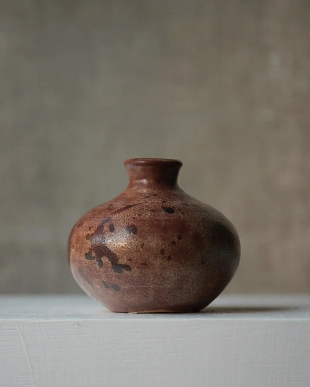 A small rounded stoneware vessel with a short neck, glazed in deep russet tones with dark speckling and patches of smoky black, photographed on a white surface against a neutral background.