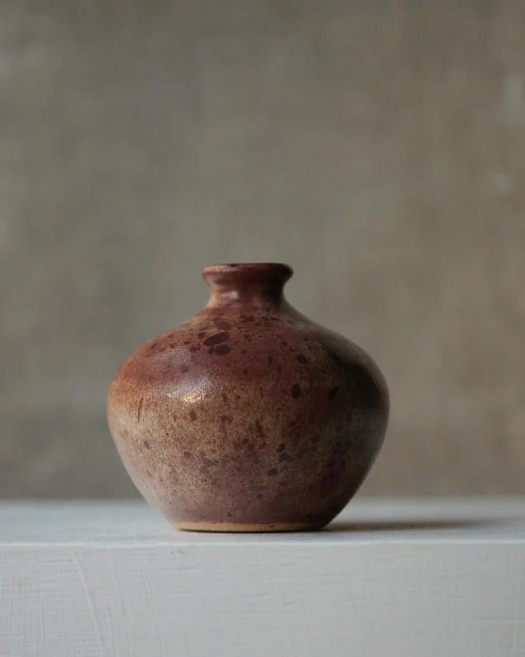 A small stoneware vase with a warm brown glaze mottled with darker patches and scattered black speckles, shown on a white plinth with a soft beige backdrop.