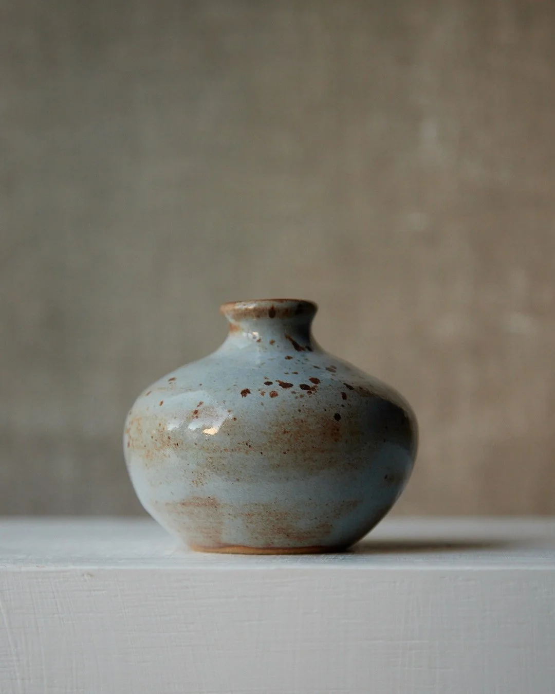 A small rounded stoneware vase with a short neck, finished in soft blue glaze with concentrated rust-toned freckles on the shoulder, shown on a white surface with a muted beige backdrop.