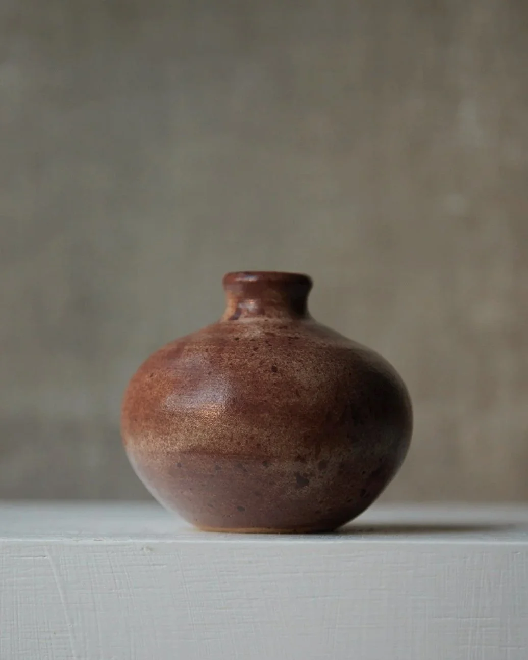 A compact rounded stoneware vessel glazed in warm earthy browns with lighter speckling and a soft matte finish, photographed against a muted neutral background.