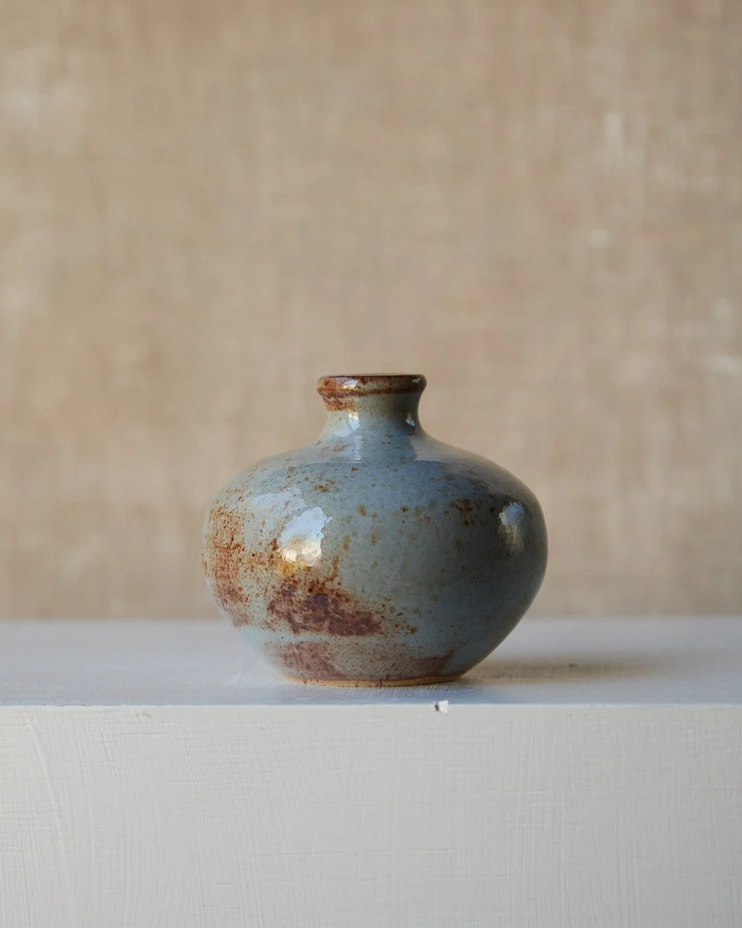 A small stoneware vessel with a short neck and rounded form, glazed in pale blue with prominent rust-toned patches on one side, shown against a softly textured neutral background.