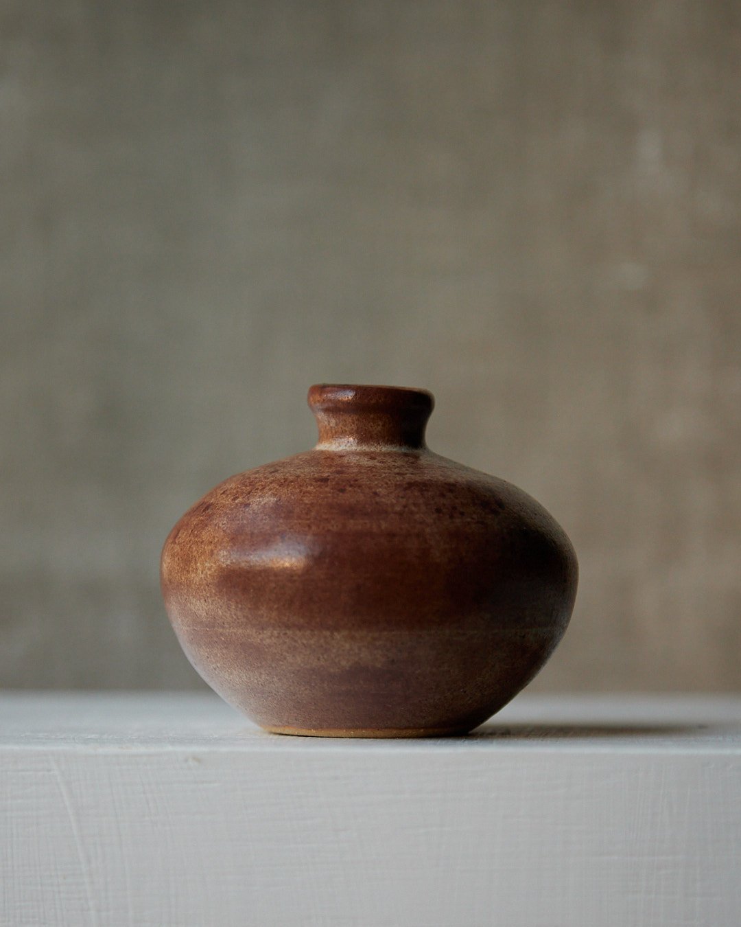 A small stoneware vase with a softly rounded form, glazed in deep brown with lighter areas along the shoulder and fine iron speckles, photographed on a white surface with a beige backdrop.