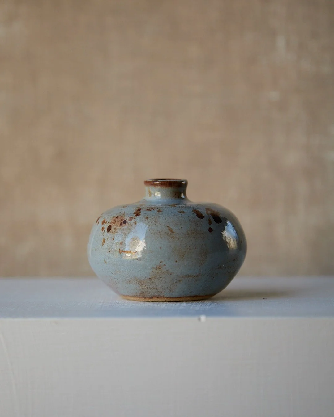A small blue-glazed stoneware vessel with a wide rounded body and short neck, featuring scattered warm brown speckles near the rim, set on a white surface against a neutral background.