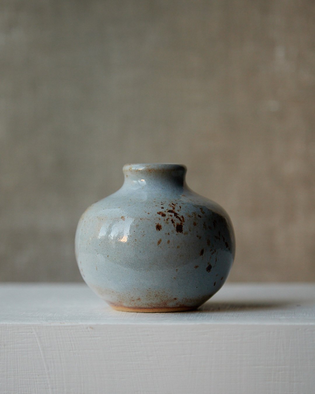 A compact rounded stoneware vessel with a narrow neck, glazed in light blue with fine iron speckles and warm clay tones visible near the base, photographed against a neutral textile background.