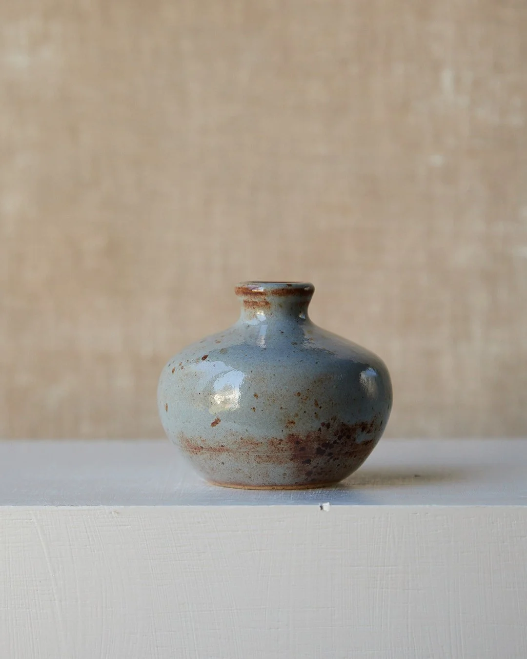 A small rounded stoneware vase finished in soft blue glaze with areas of warm rust-colored mottling near the lower half, photographed on a white pedestal against a warm beige backdrop.