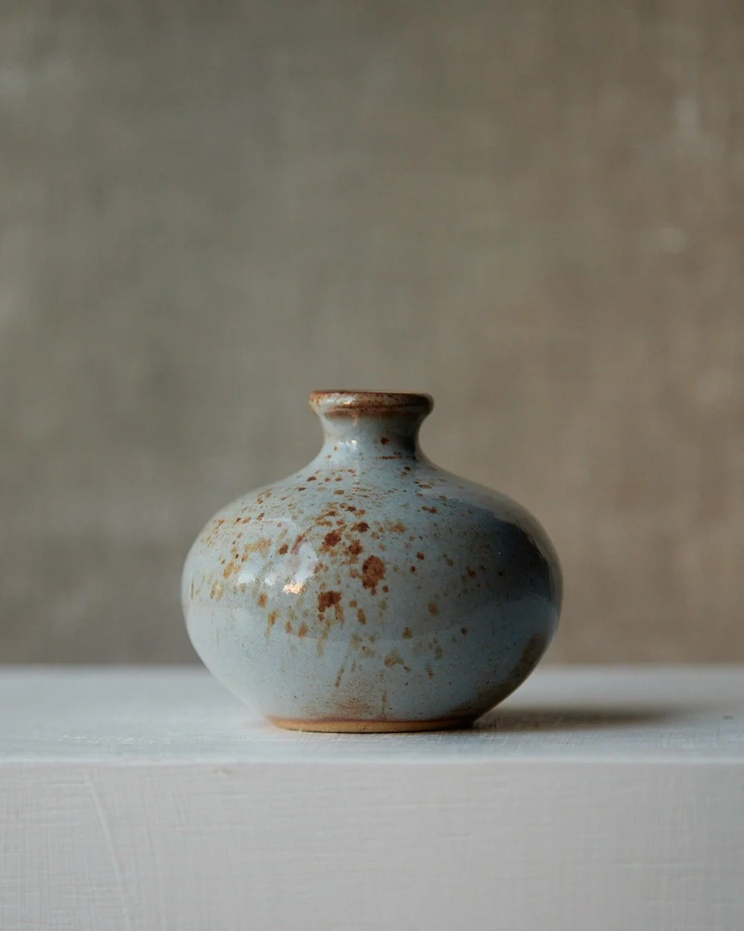 A small round stoneware vessel with a narrow neck, glazed in pale misty blue with warm rust-colored speckles, photographed on a white surface against a neutral background.