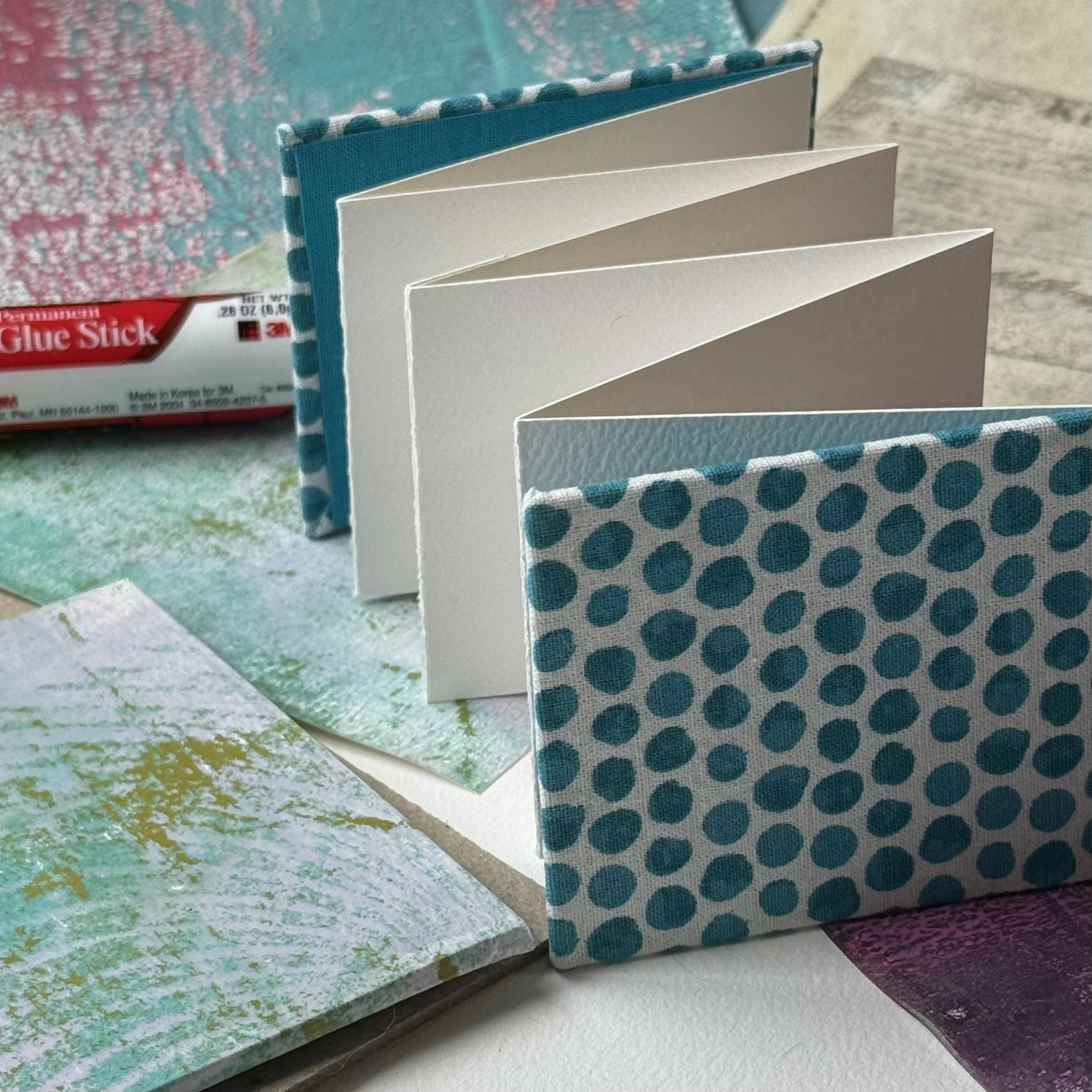 Open blank watercolor notebook with a polka dot cloth cover, placed on a textured surface with an art project and glue stick in the background.