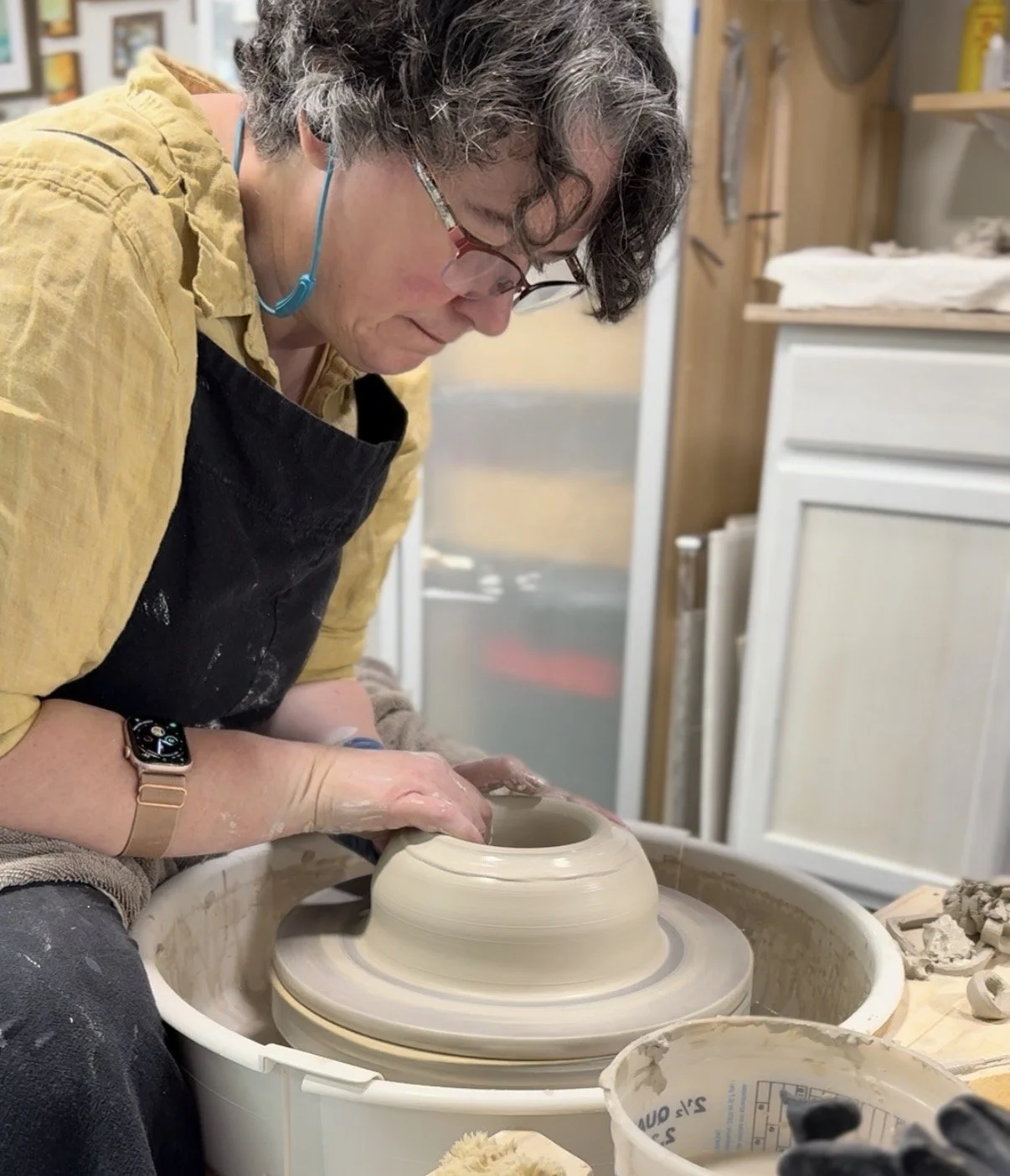 A person with glasses and a yellow shirt working on a pottery wheel, shaping a ceramic vase or bowl, in a pottery studio.