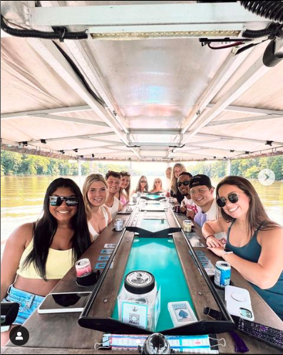 Group of friends enjoying a boat ride at a long table on a boat, with drinks and mobile phones, surrounded by water and greenery.
