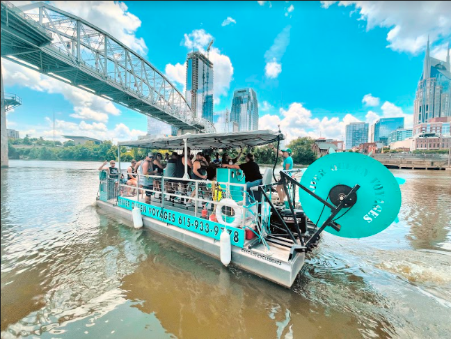 A sightseeing boat cruises with passengers on the Cumberland River in Nashville, with a bridge and skyscrapers in the background, and a large turquoise paddlewheel.