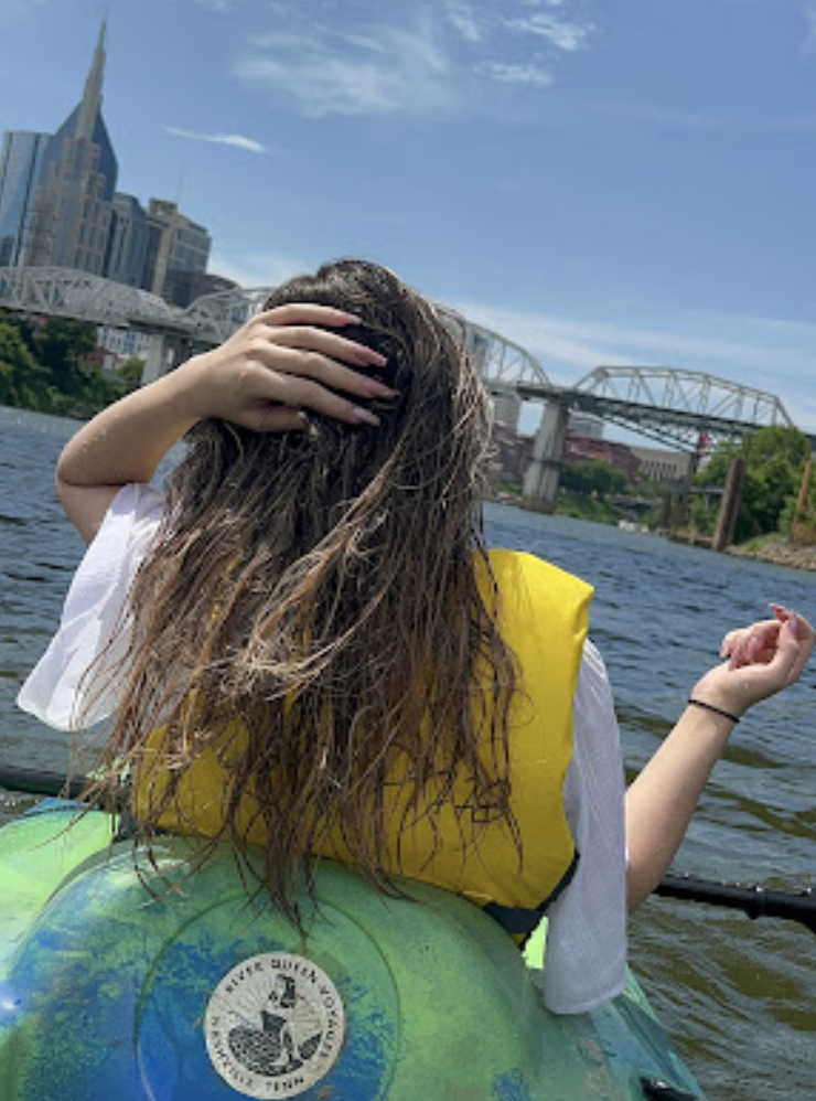 Person with long hair sitting in a kayak on a river, wearing a yellow life jacket, with a city skyline and a bridge in the background.