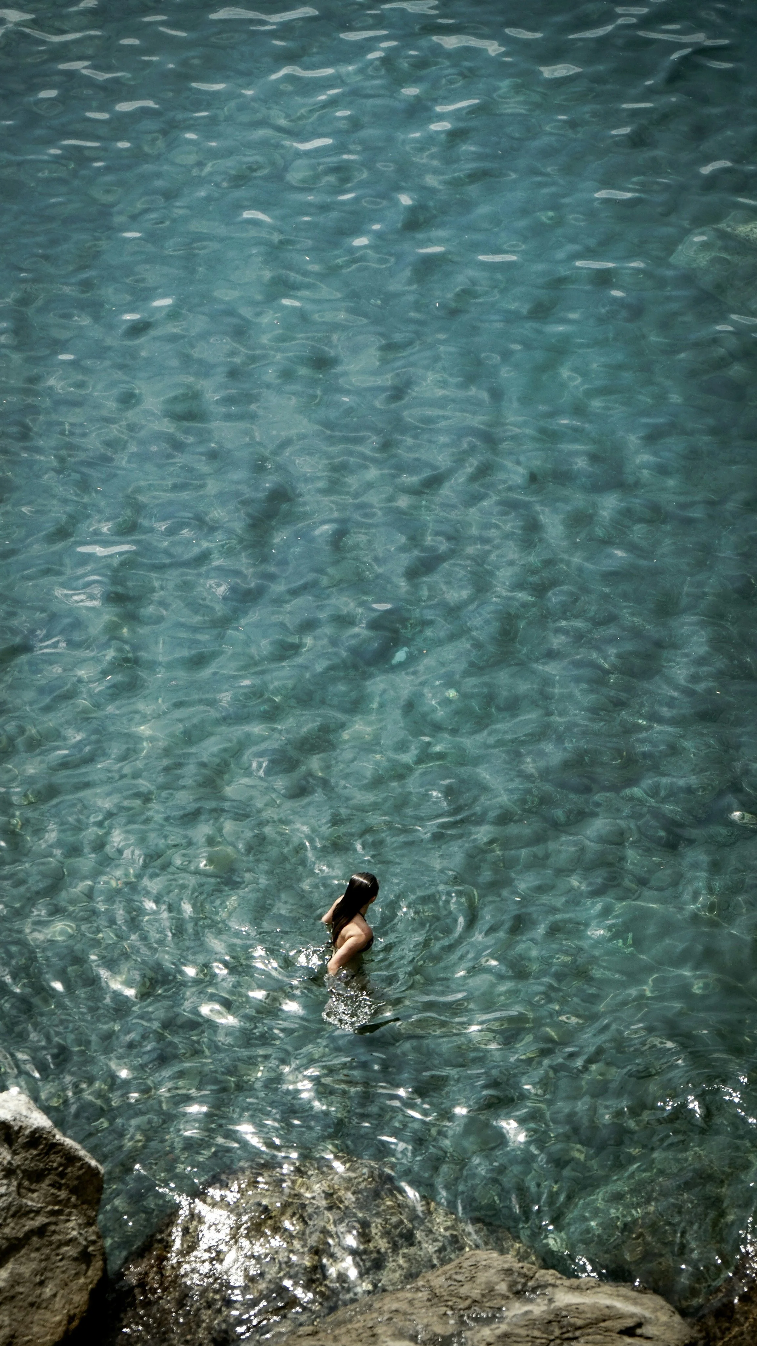 A person with long dark hair swimming in clear blue water near rocks.
