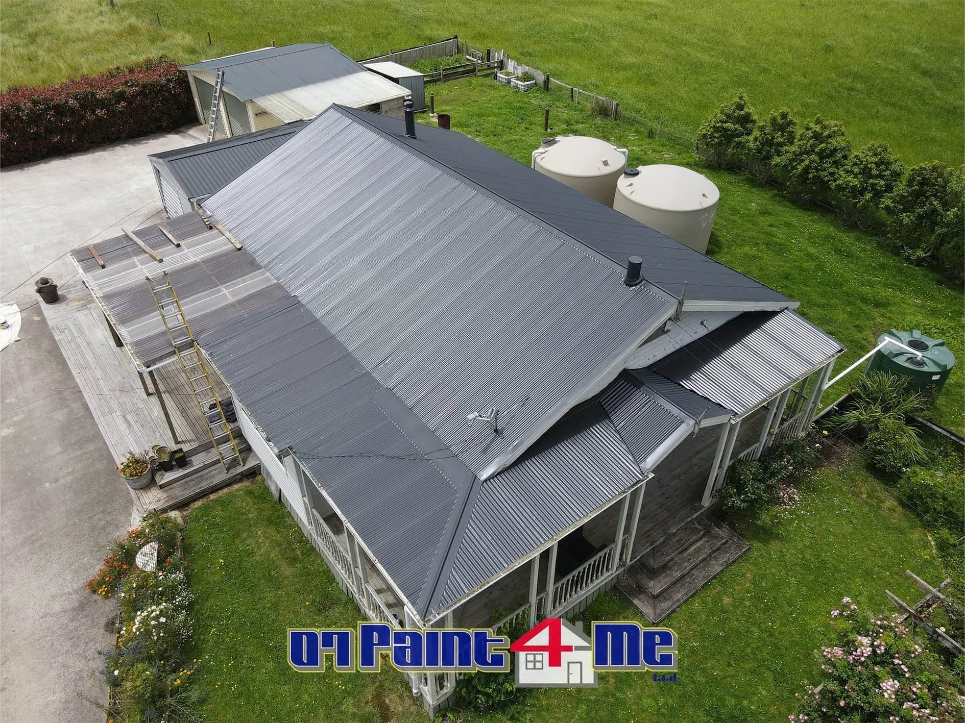 Aerial view of a farmhouse with a freshly painted metal roof surrounded by a green yard, water tanks, and a shed.