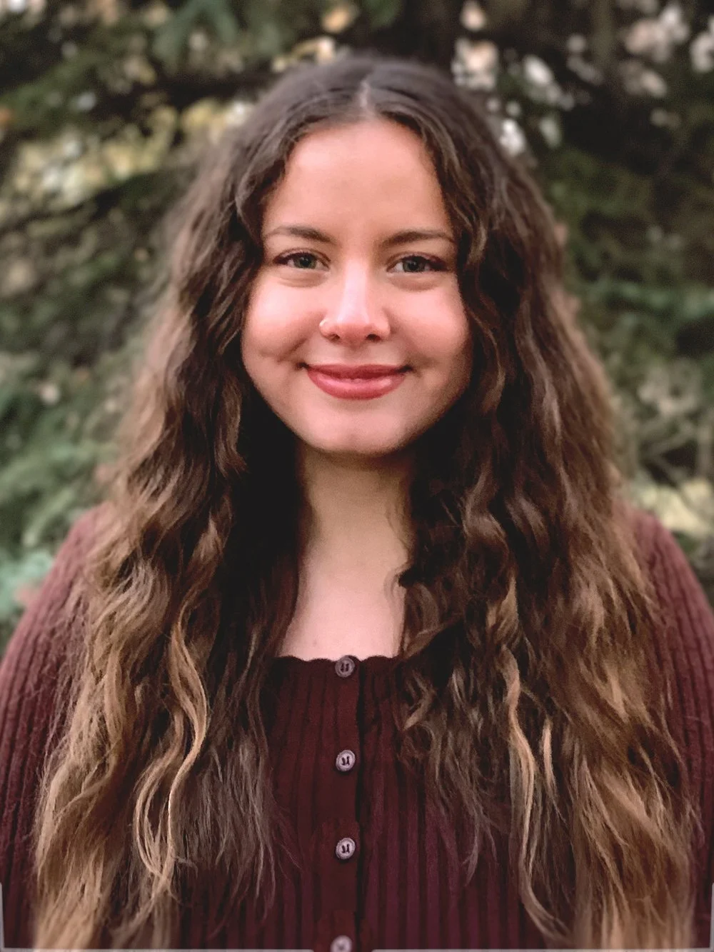 An image of Luiza de Queiroz - Registered Massage Therapist (RMT) -  a young woman with long, wavy brown hair smiling outdoors with a blurred green and white background.