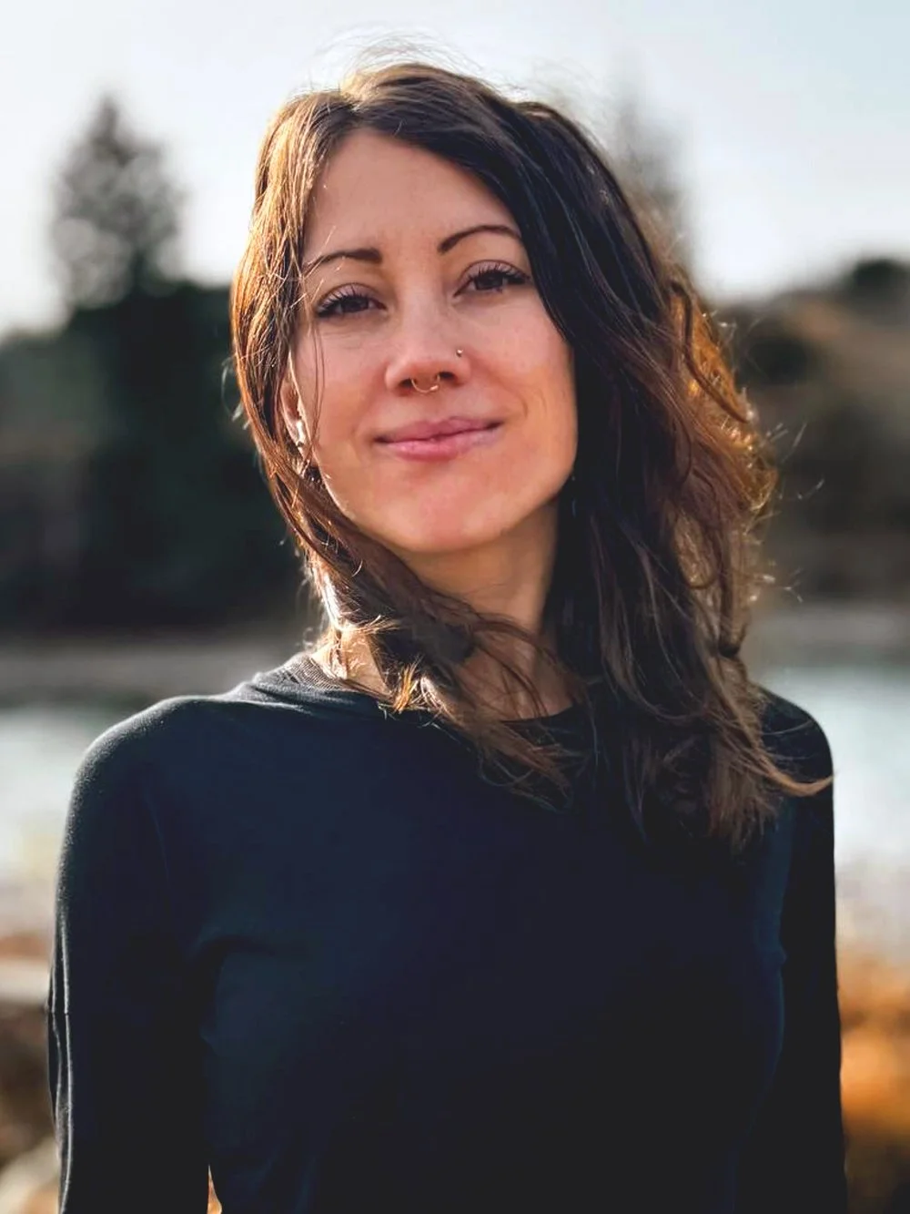 Cassandra Musser Lamoureux is a Registered Massage Therapist (RMT)  at Omega Massage + Wellness with shoulder-length brown hair, wearing a black top, standing outdoors with a blurred background of trees and water, smiling gently at the camera.