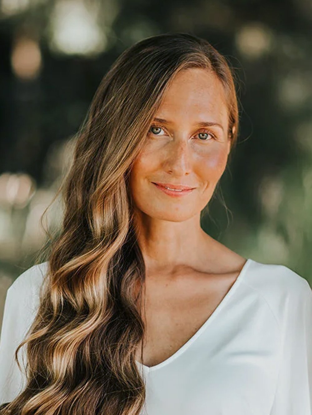 Image of Mellisha Fehr, Founder, Director and Registered Massage Therapist (RMT) at Omega Wellness + Massage. Mellisha has long wavy brown hair and blue eyes, is wearing a white top, smiling outdoors with blurred trees in the background.