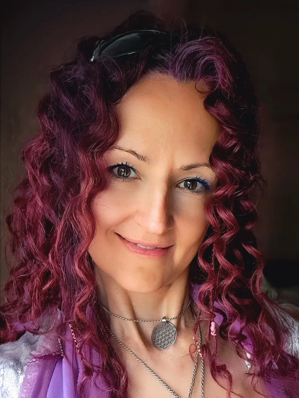 Jolene Tse is a Registered Massage Therapist (RMT) and Energy Practictioner at Omega Massage + Wellness. She has curly purple hair, light skin, and blue eyes smiling at the camera, wearing layered necklaces and glasses on her head.