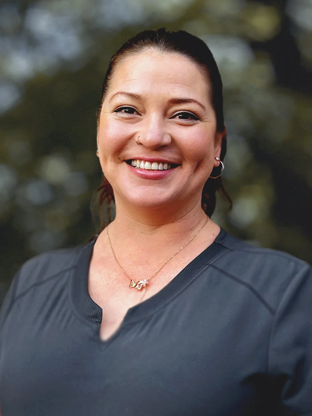 Carole Cadle is a Registered Massage Therapist (RMT)  at Omega Massage + Wellness . She is smiling with dark hair and earrings, wearing a black top and a gold necklace, outdoors with blurred green foliage in the background.