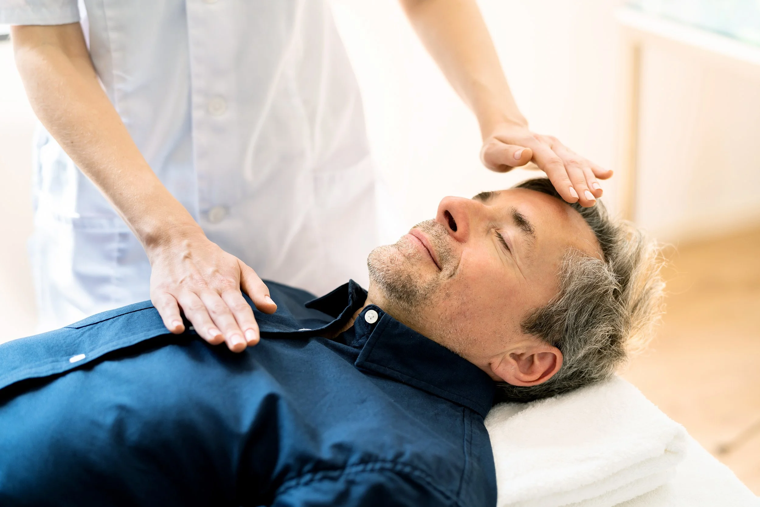 Smiling Caucasian man lying face up with massage therapist's hands hovering over forehead and chest