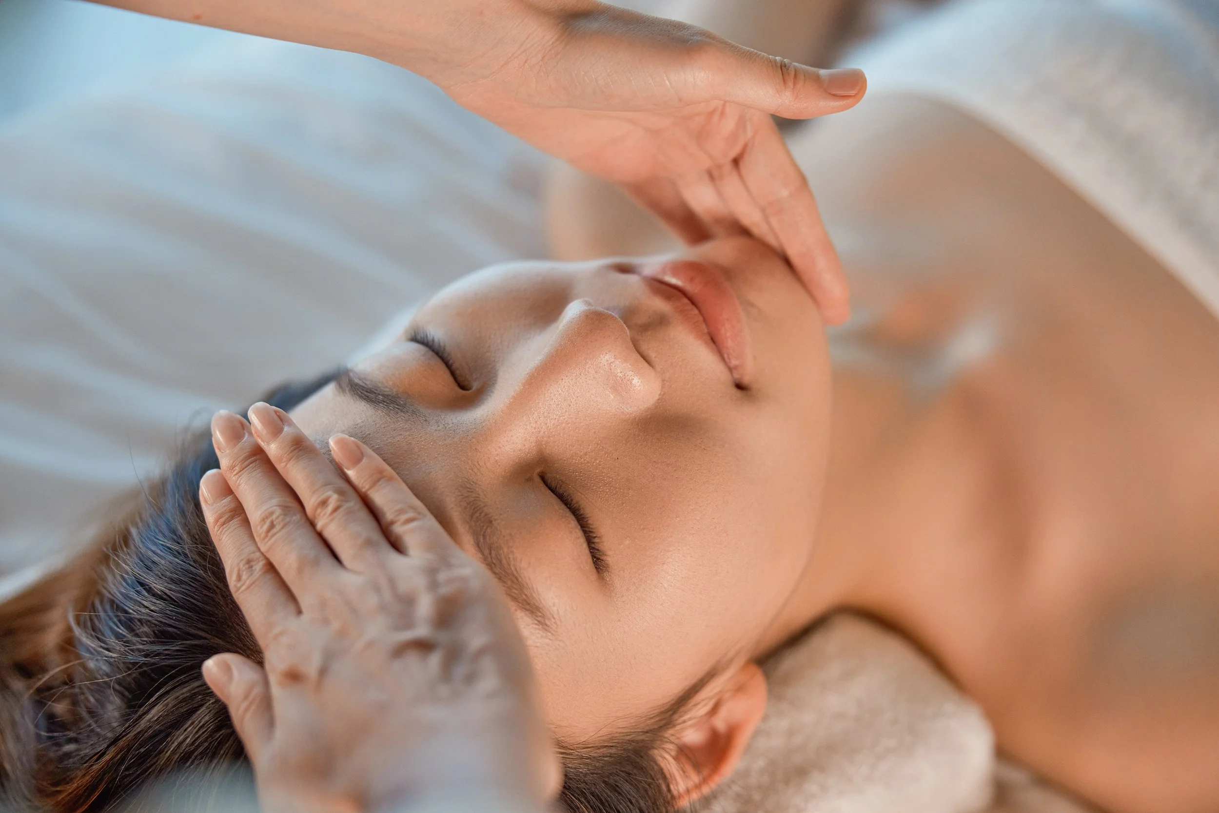 Smiling Asian woman lying face up with massage therapist's hands on forehead and under chin providing support