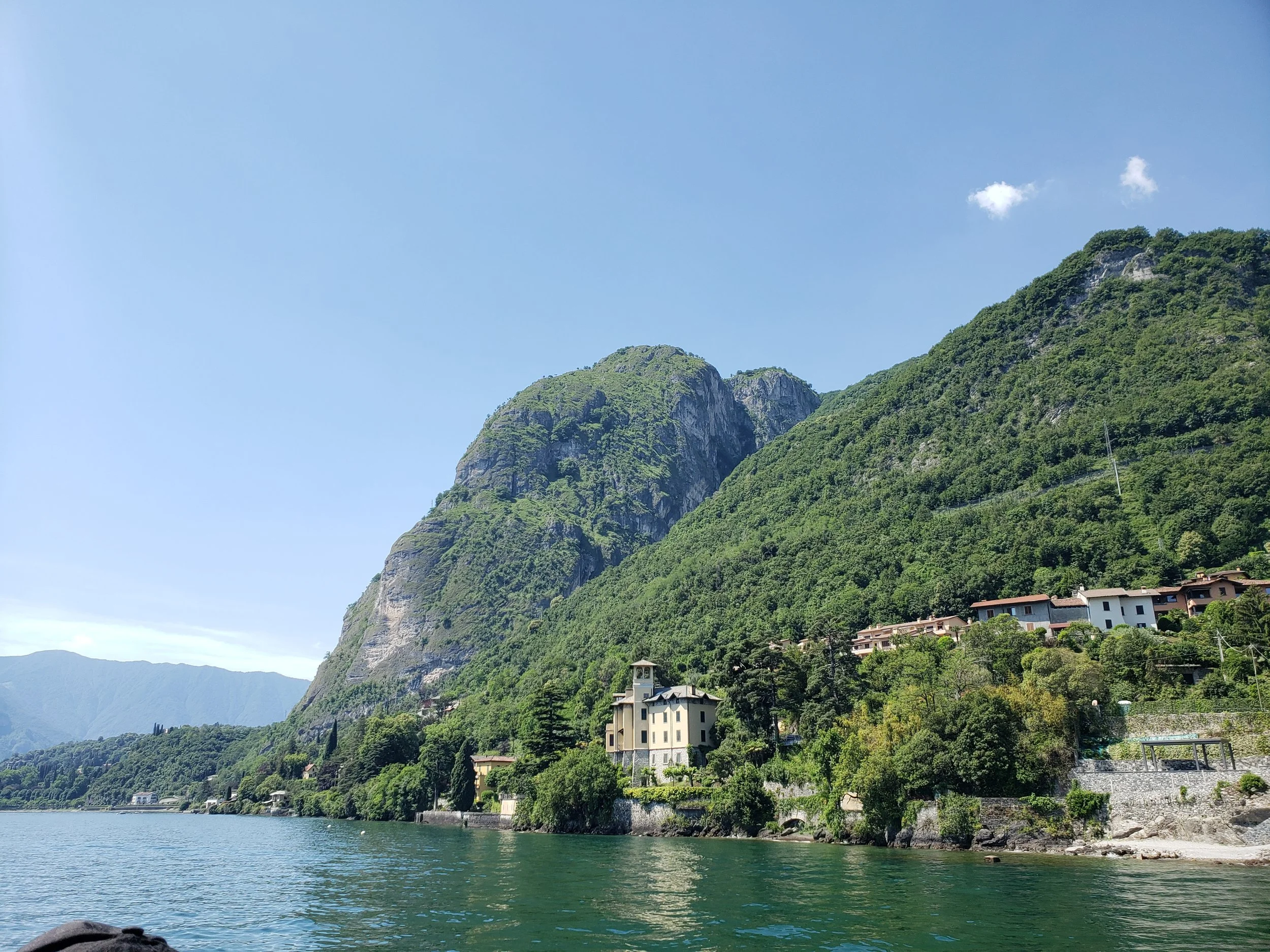 A scenic view of a lake with clear, calm water in the foreground, bordered by a rocky shoreline. In the background, lush green mountains rise steeply, dotted with trees and some buildings near the base of the mountains. The sky above is clear and blue with a few small clouds.